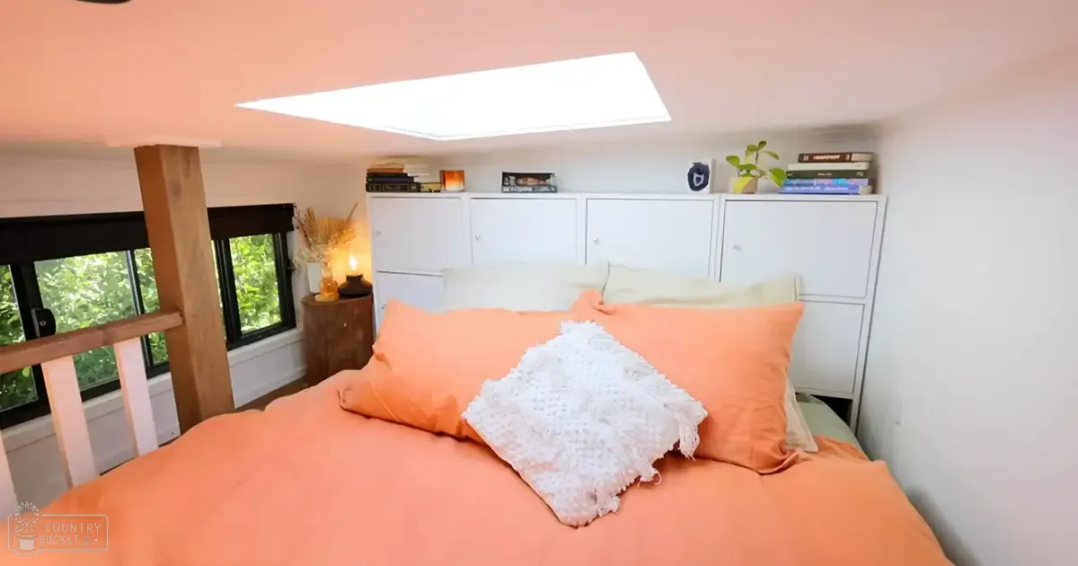 A cozy bedroom with orange bedding, white pillows, wooden nightstand holding dried grasses in a vase and a small lamp emitting light, white storage cabinets containing books and plants above the bed, natural light coming through windows with black frames showing greenery outside.