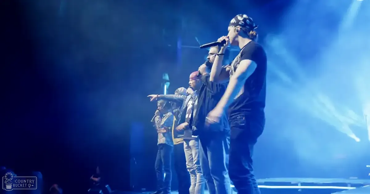 A group performing on a blue-lit stage; adult males in casual attire hold microphones during what appears to be a concert.