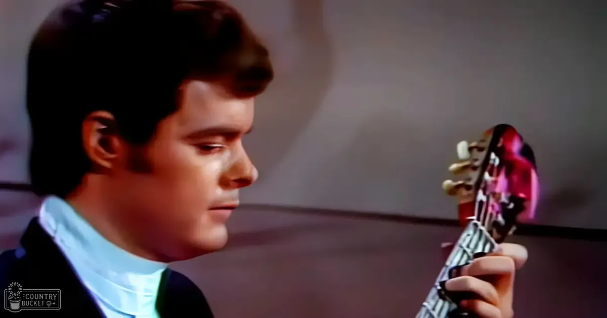 A young adult male in a tailored dark blazer and light blue collared shirt holding a red-body electric guitar, set against an indoor backdrop with even soft lighting.