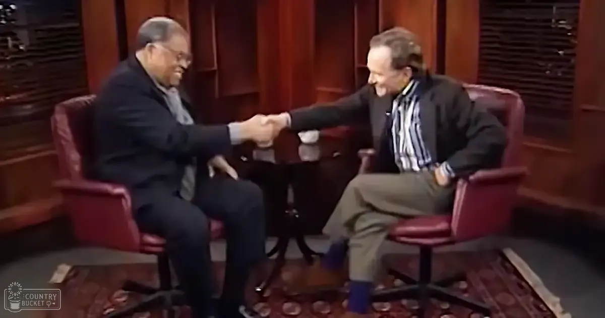 An older adult male singer in a dark suit jacket and white shirt on stage, with another middle-aged to older adult male wearing a striped button-down shirt and dark blazer, seated against two red leather armchairs in a late 20th-century talk show set.