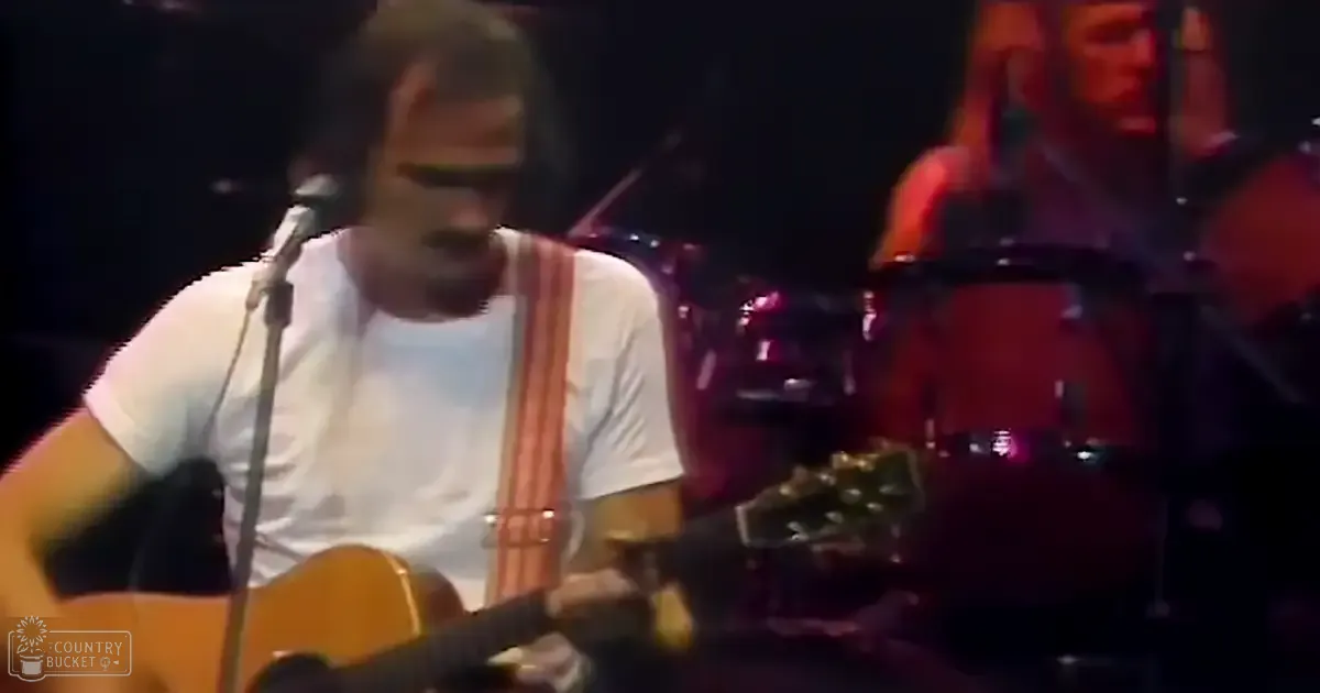 An adult male musician playing an acoustic guitar with a white t-shirt and dark pants, surrounded by stage lighting and musical equipment in a concert hall setting.
