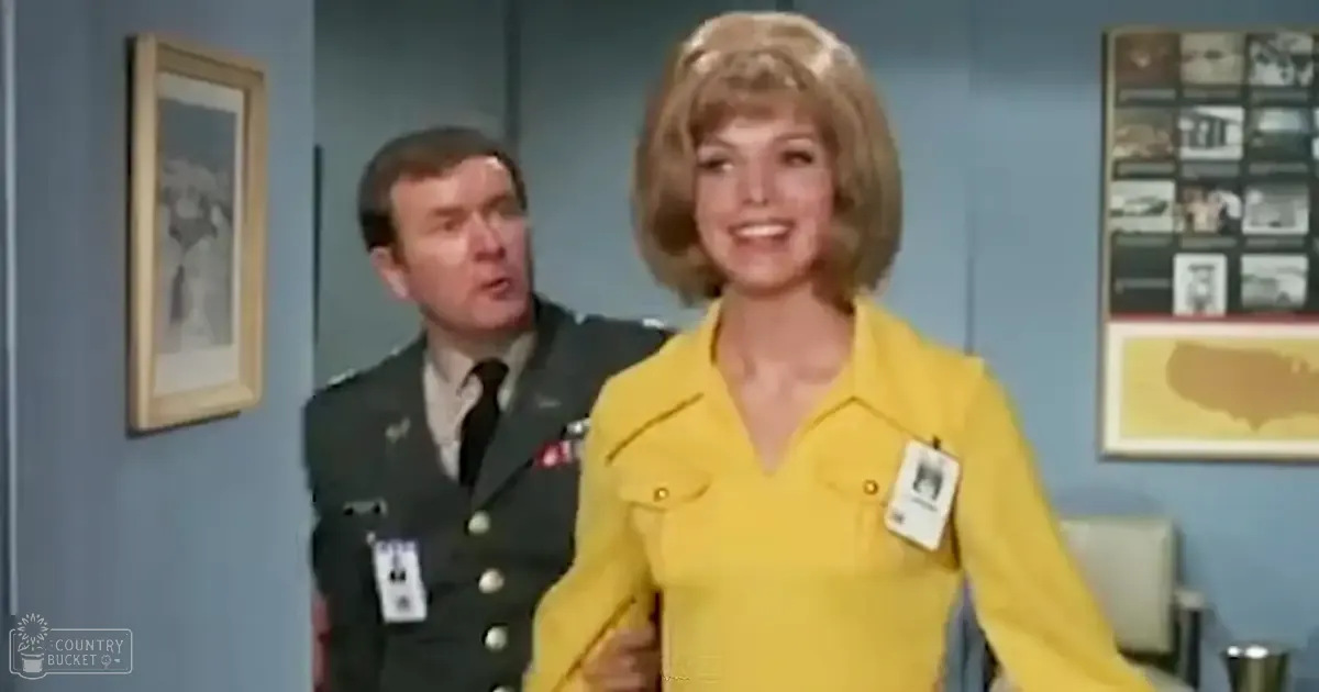 A woman in her adult years wearing a mid-20th century yellow button-up shirt, standing indoors with an adult male in a dark military-style uniform. The setting appears to be an office environment with framed pictures and wall decorations.