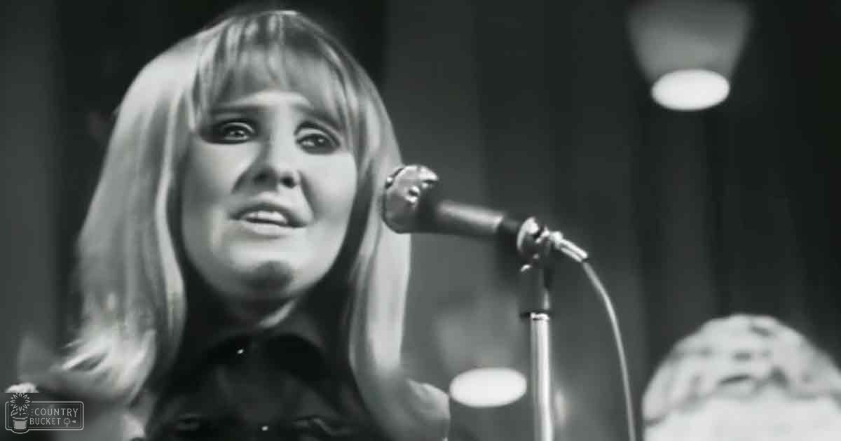 A young adult female performer wearing a dark-colored garment with long sleeves and high neckline stands before a microphone at an indoor event, possibly a concert or recording session.