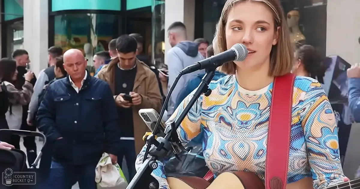 A young adult female singer performs outdoors near a shopping area, wearing a patterned blue and yellow top with red guitar strap; a man walks by carrying a stroller or baby carrier.