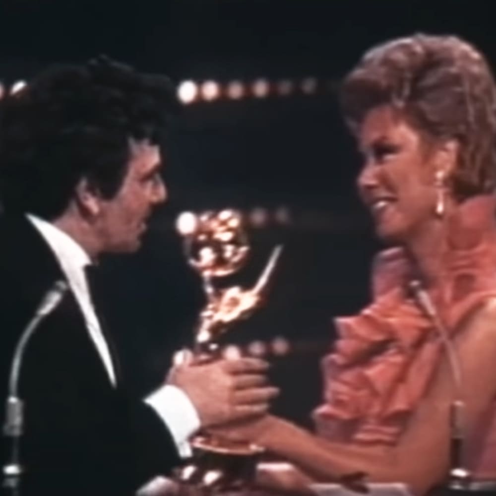 An awards ceremony scene with a male adult singer in a black tuxedo and a female middle-aged to older adult wearing a pink ruffled dress with a low-cut neckline, both holding a trophy on stage under circular lights.