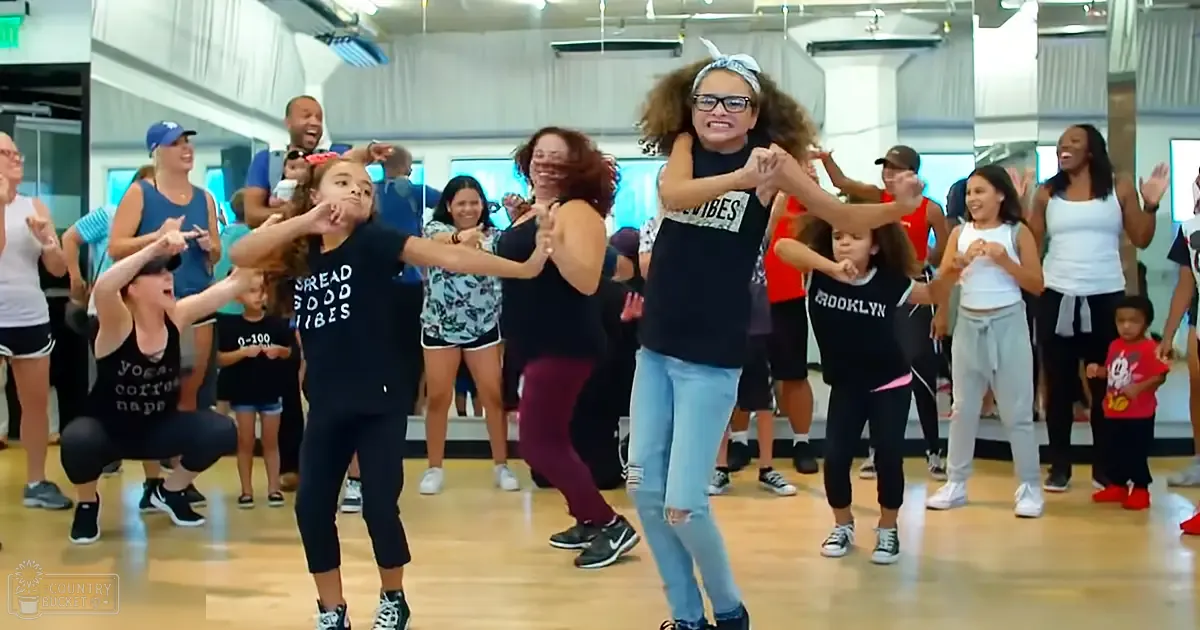 Children in a group dance class wearing casual attire in an indoor studio with mirrors, wooden floors, and fitness equipment.