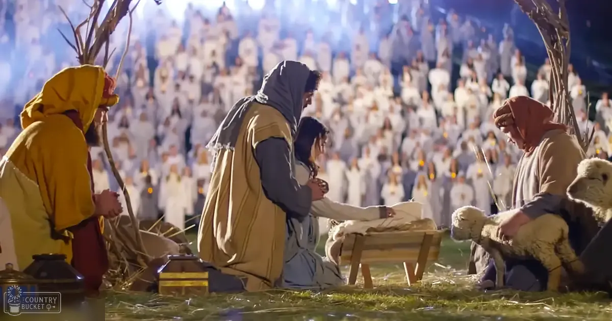 A nativity scene indoors, likely in a church or religious gathering place, with two adults dressed as Mary and Joseph kneeling beside the manger, surrounded by other figures and an adult holding a sheep. The setting includes wooden furniture like a manger and a small crate, with a large crowd of people dressed in white in the background.