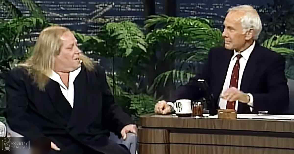 Two adults in formal attire seated at a desk with mugs and a greenery background resembling an indoor studio environment.