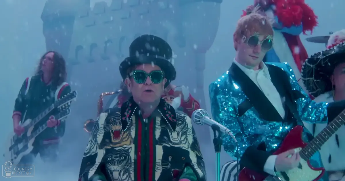 Adult males performing at a winter outdoor concert venue with foggy background, stone structure resembling a castle in distance; blue sequined blazer, black top hat, sunglasses with reflective lenses.