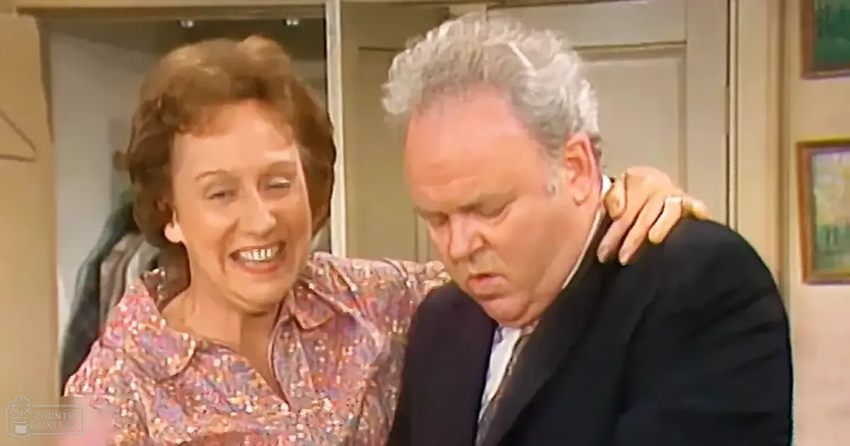 Middle-aged woman wearing a floral blouse indoors alongside a middle-aged man in formal attire; domestic interior with framed artwork visible.