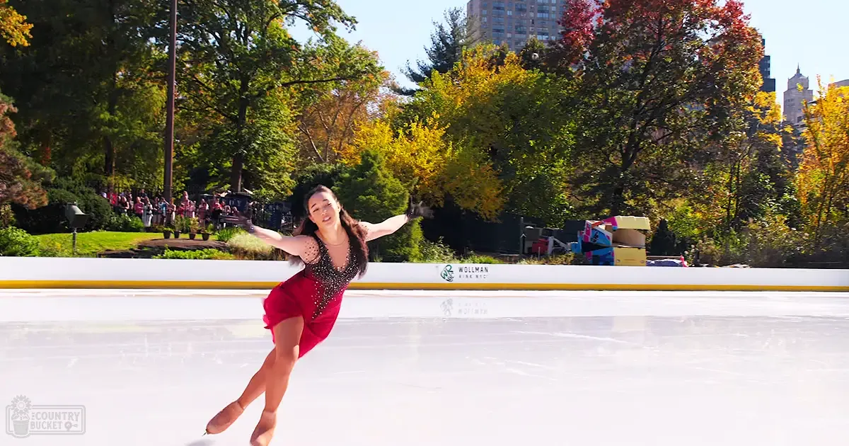 A young adult female figure skating on an ice rink in Central Park, surrounded by trees and a crowd of spectators.