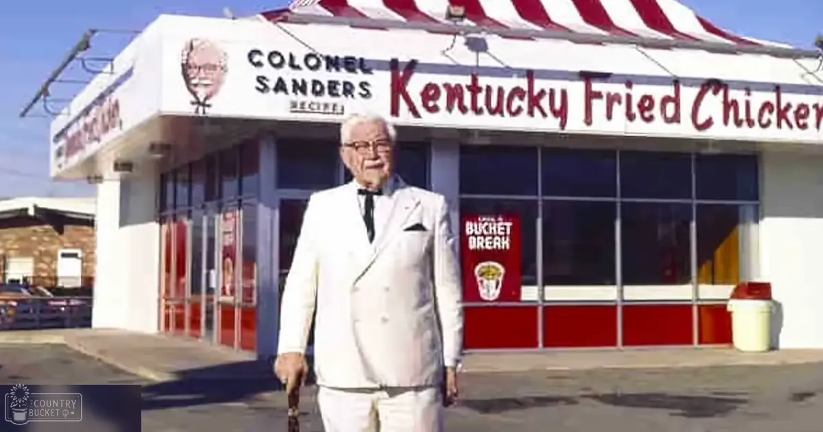 Colonel Sanders in a white suit jacket with black lapels and buttons stands outside a Kentucky Fried Chicken restaurant.