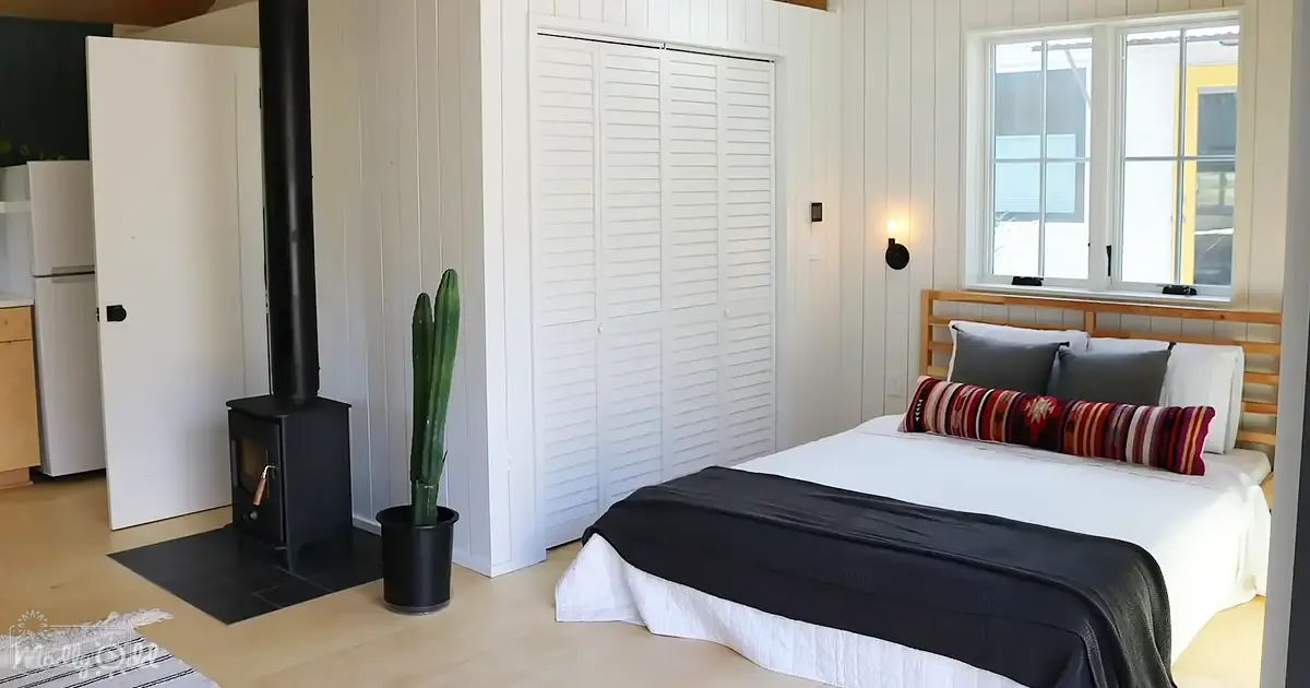 A modern bedroom with white bedding and black throw blanket, wooden flooring, large window allowing natural light.
