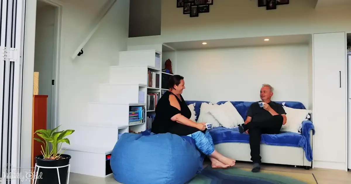 Two adults converse in a cozy living room with natural light, with blue bean bag chair, white sofa and bookshelf filled with books.