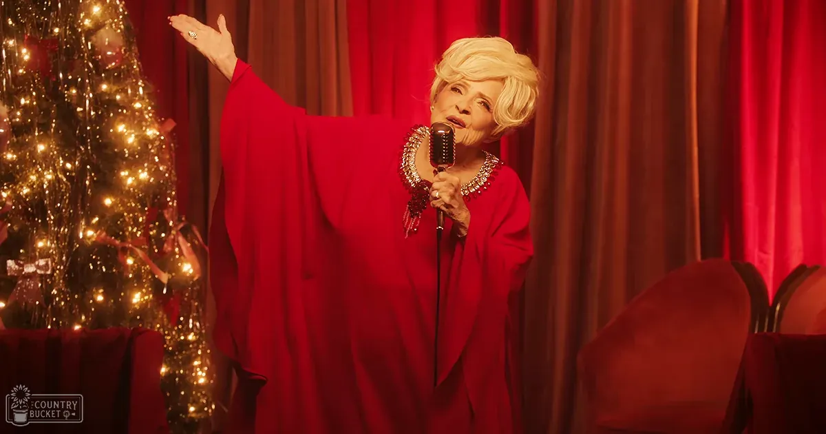 An older adult female in an elegant red robe, possibly singing on stage with a Christmas tree and red curtains behind her.
