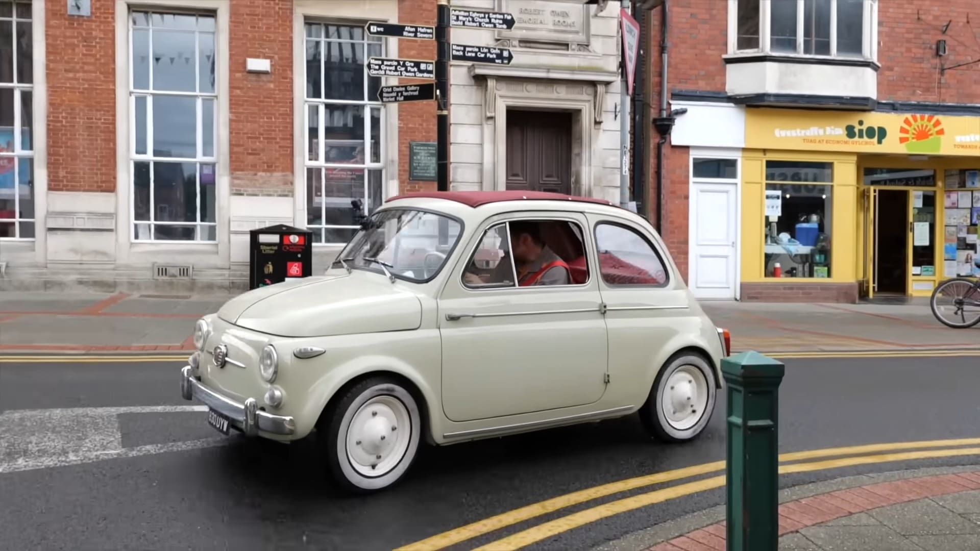 An adult male in casual attire stands beside a vintage Fiat 500 car parked on an urban street near a tomato shop with a sun logo.