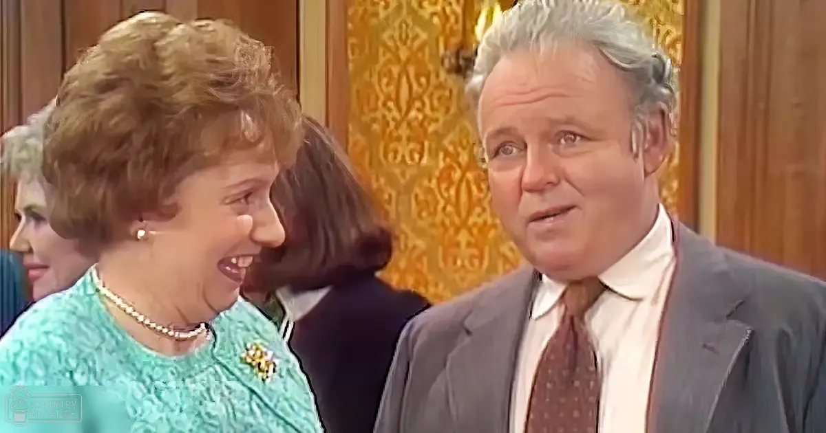 Middle-aged man in a dark gray suit jacket with pearl accessories, standing next to a middle-aged woman in a light teal floral dress with ruffled texture and gold wallpaper backdrop.