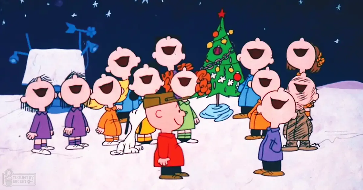 Children in colorful holiday outfits gather outdoors at night against a backdrop of a decorated Christmas tree and snow-covered house with a windmill, under a starry sky.