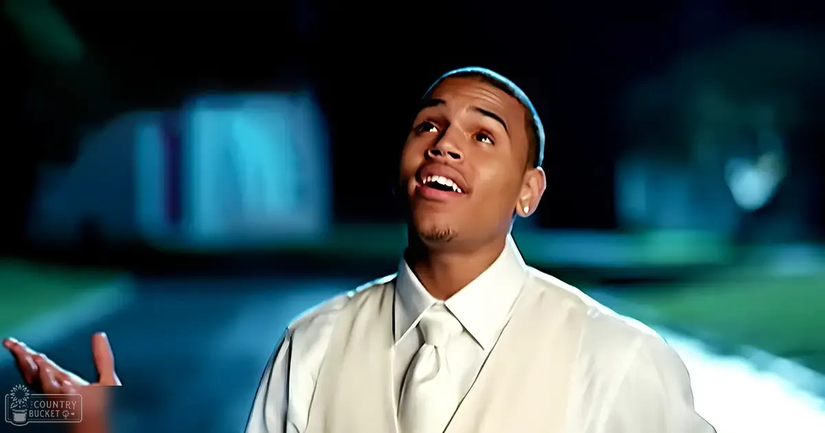 A young adult male stands outdoors at night, wearing a white dress shirt with a white tie, small earring visible. The setting appears to be a park or garden area illuminated by artificial light.
