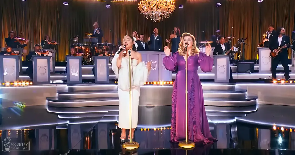 Musical performance scene with two adult females in a formal indoor setting, with a chandelier and musicians playing violins, drums, saxophones, and guitars.