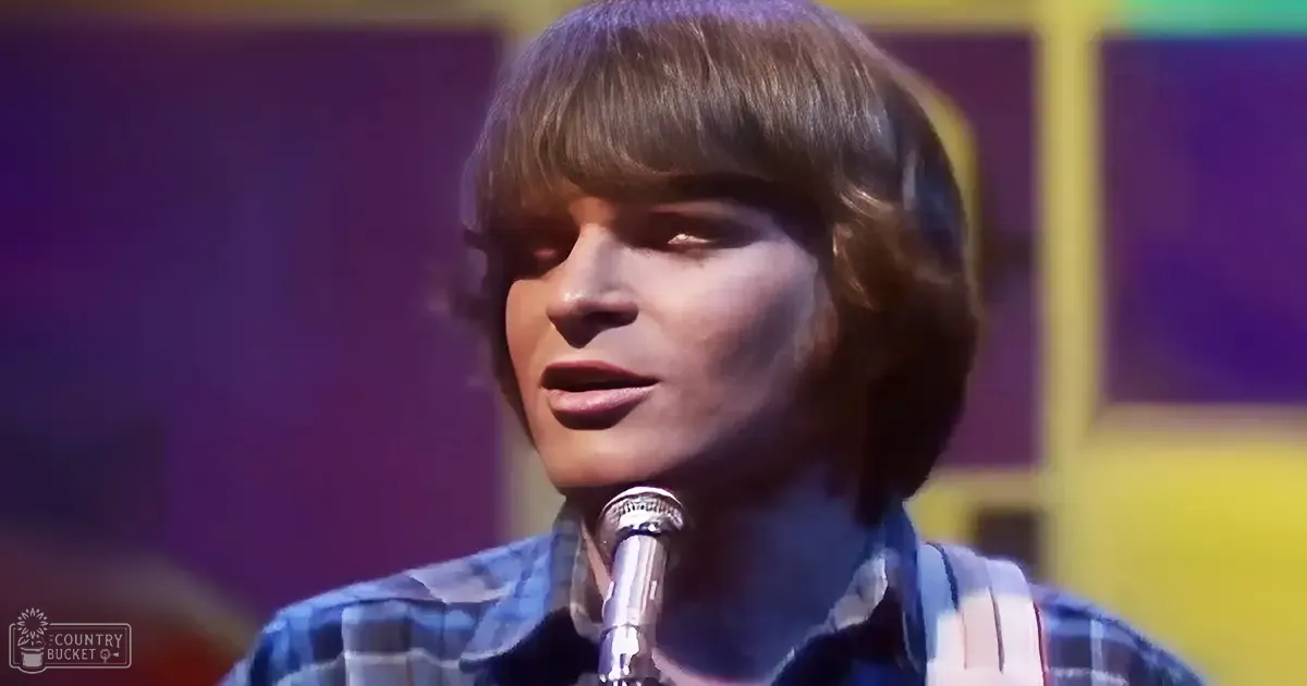 A young adult male singer wearing a classic mid-20th-century plaid shirt performs into a microphone at an indoor venue with bright lighting and a purple backdrop.