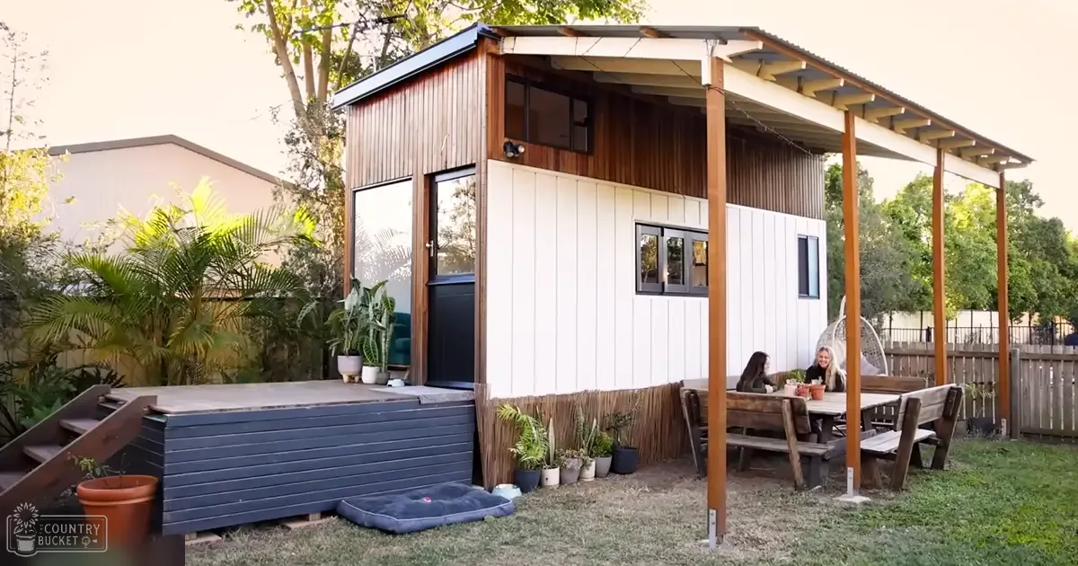 A modern tiny house with white siding and wooden accents in a residential backyard setting, surrounded by outdoor furniture and potted plants under a covered porch area.