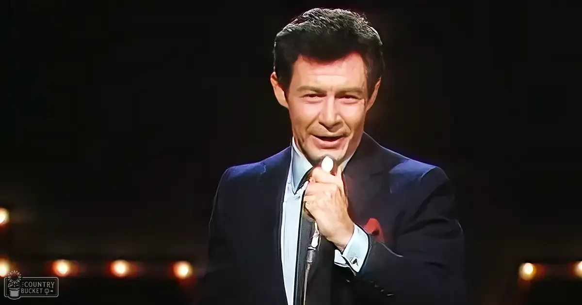 A middle-aged adult male in a dark blue suit jacket with white shirt and tie performing on stage under dim, warm spotlights.