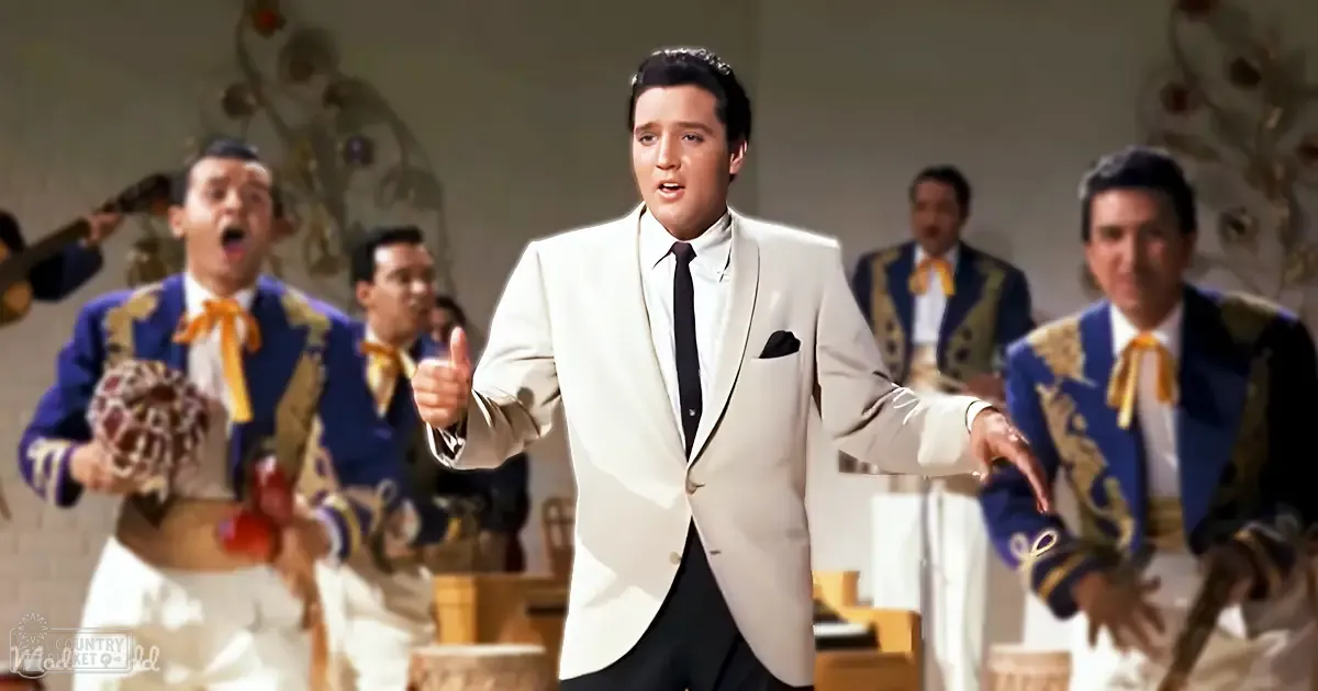 An adult male in a white suit jacket with a black pocket square and dark tie, standing indoors. Behind him are adults wearing blue jackets with gold embroidery and orange bow ties, likely part of a mariachi band or performance group.