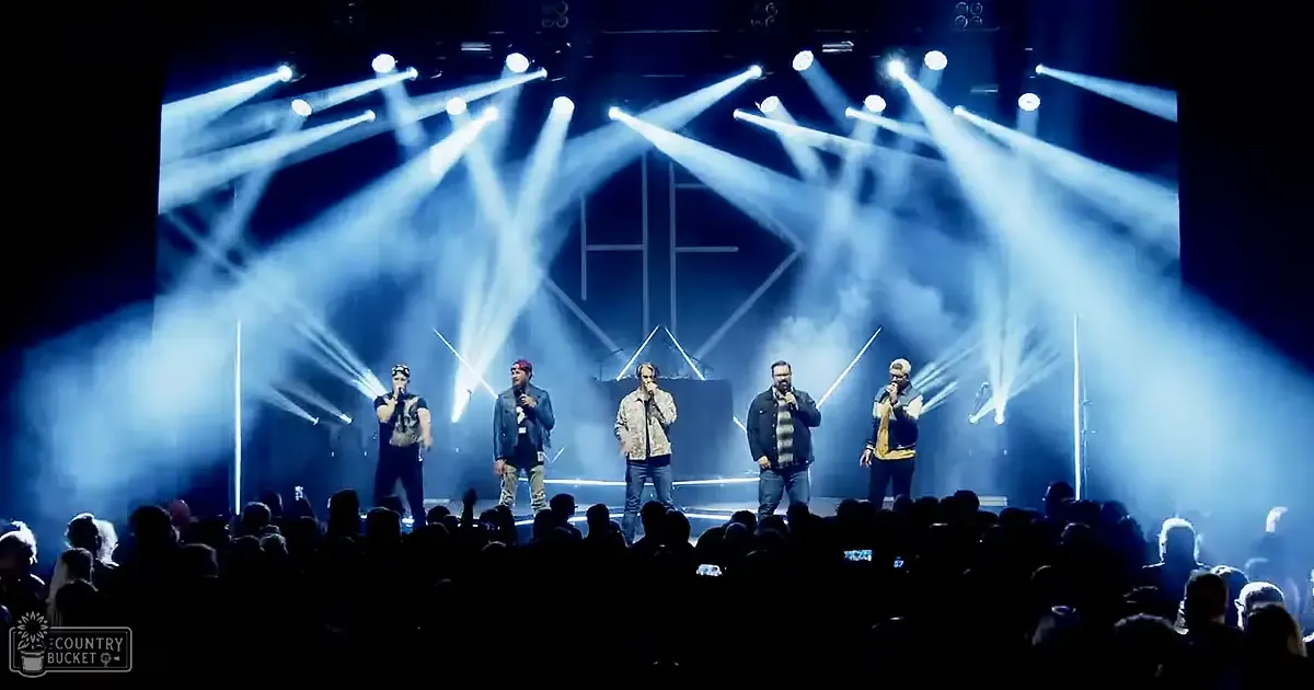 An indoor concert venue with performers on stage, bright stage lighting, and a dark audience silhouetted against the backdrop.