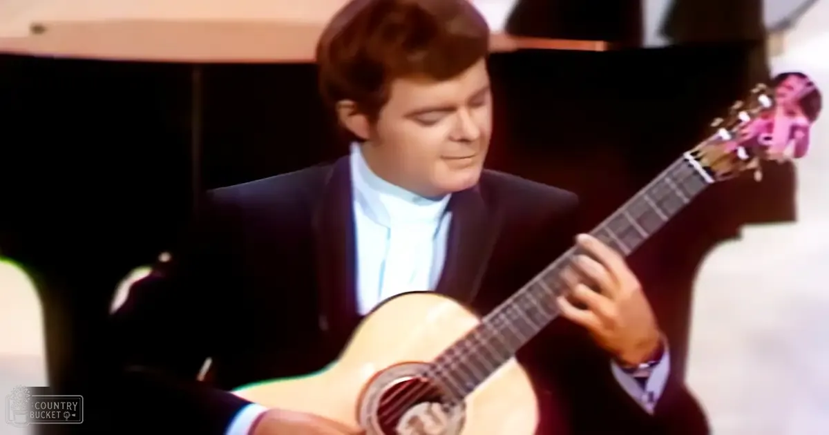 A young adult male in a dark suit jacket and white shirt with a bow tie is playing an acoustic guitar, set against the backdrop of what appears to be an event or performance venue.