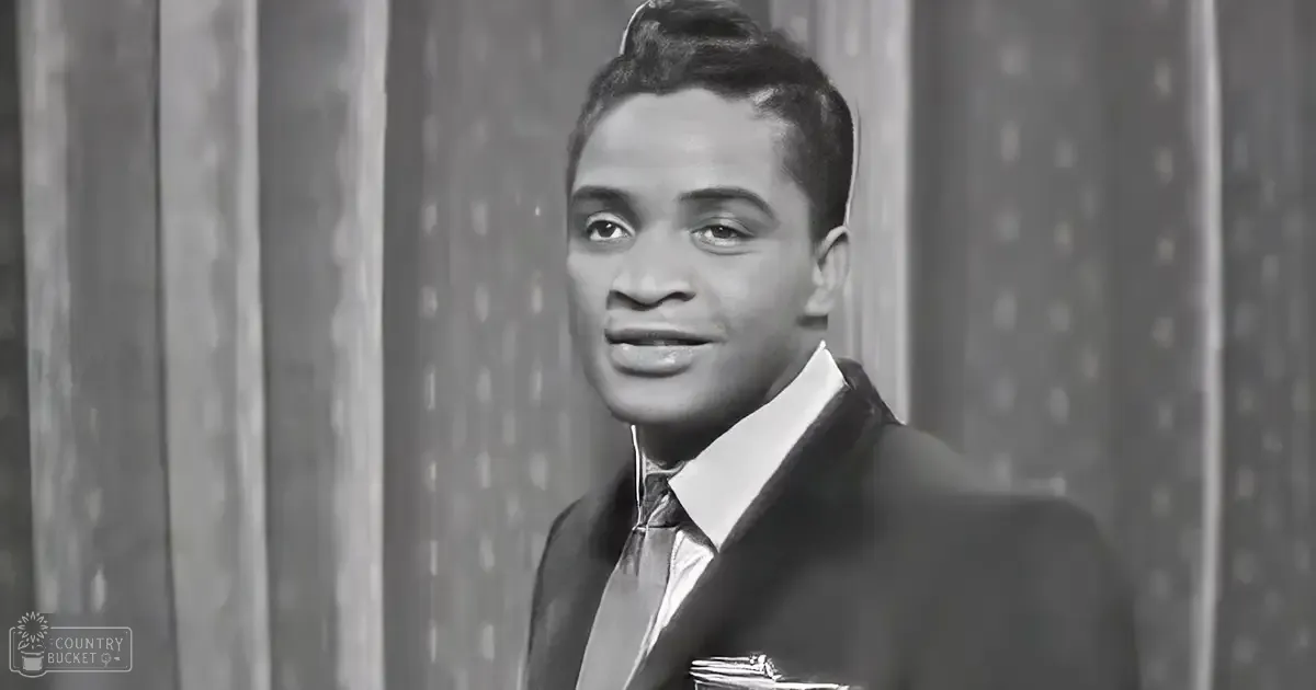 A young adult male wearing a dark suit jacket with a pocket square, set against a backdrop with vertical stripes and dots in an indoor setting.