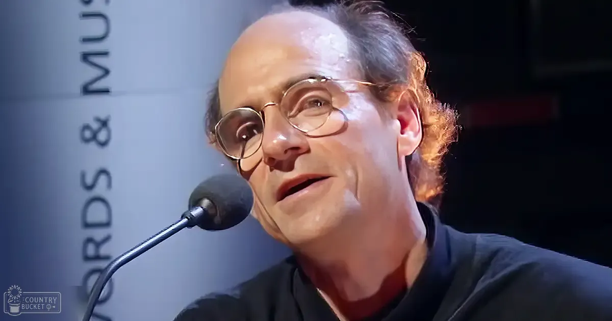 An adult male wearing a dark-colored shirt with round glasses at an indoor event focused on words and music.
