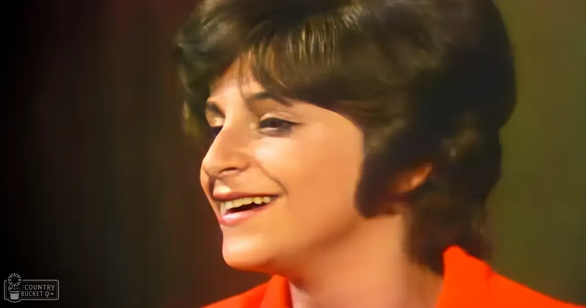 A middle-aged woman in an indoor setting, wearing a bright orange mid-20th century-style garment with high neckline and short sleeves.