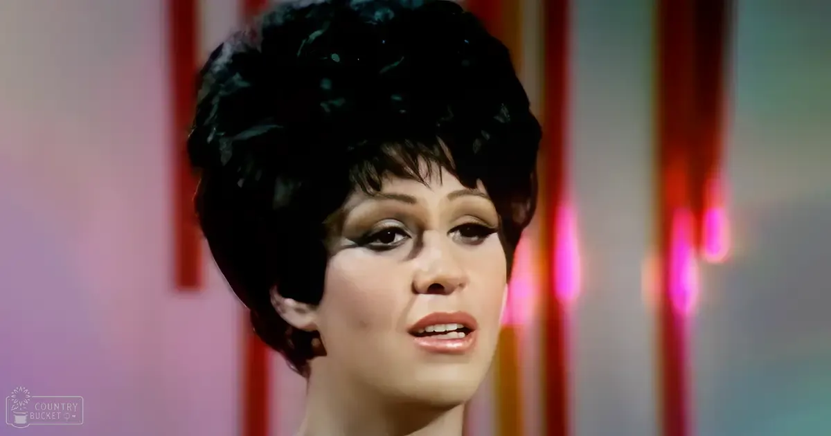 A young adult female wearing a black 1960s-style garment with high neckline and short sleeves stands against a vibrant stage-like backdrop of vertical striped fabric in an indoor performance setting.