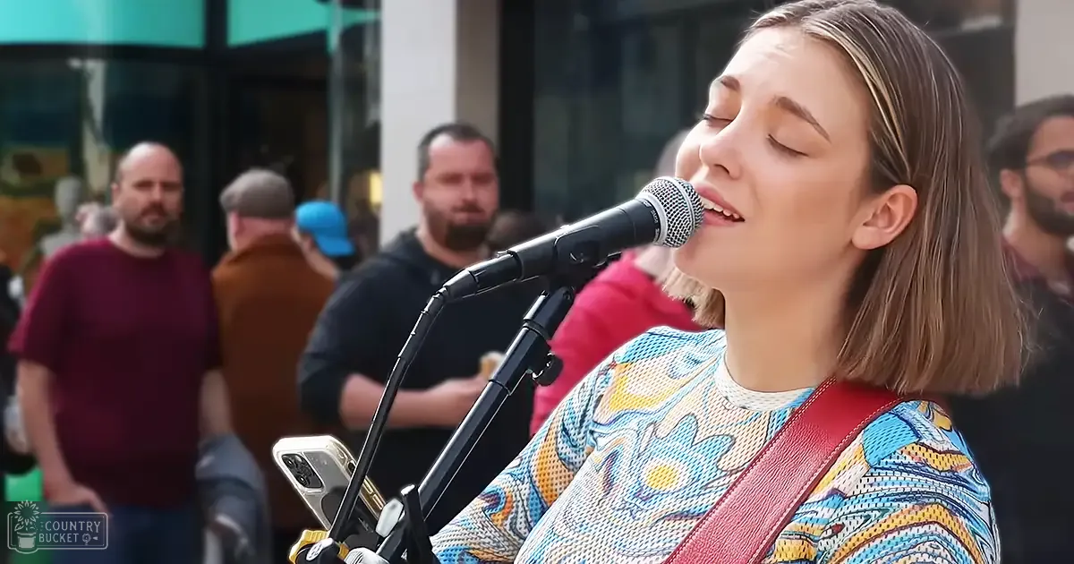 A young adult female in a colorful floral top sings outdoors, accompanied by several adults in casual attire; urban setting with microphone and smartphone visible.
