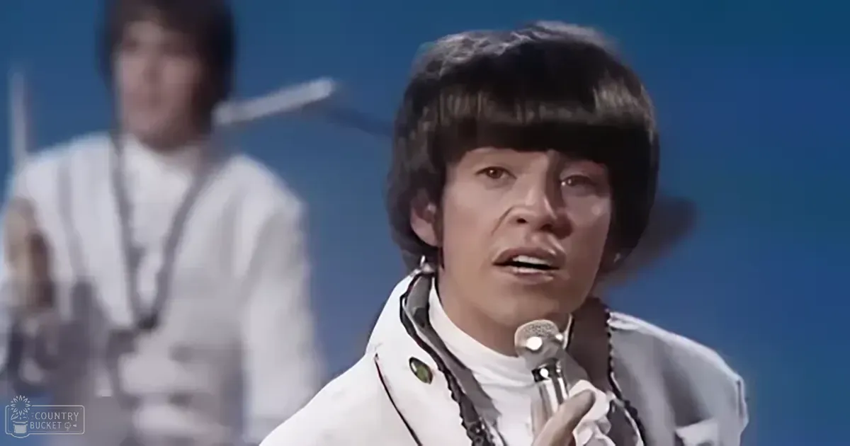 A young adult male singer wearing a white jacket with black trim, singing into a microphone on stage.