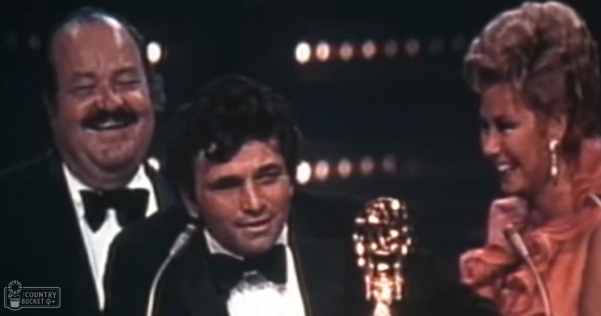 Adult male in a black tuxedo at an awards ceremony with others in formal attire, surrounded by microphones and an Oscar statue.