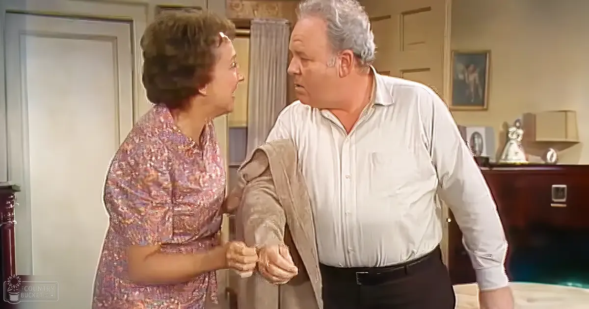 An older adult woman in a vintage dress stands indoors next to a man wearing a white shirt in a neutral-toned living room setting.