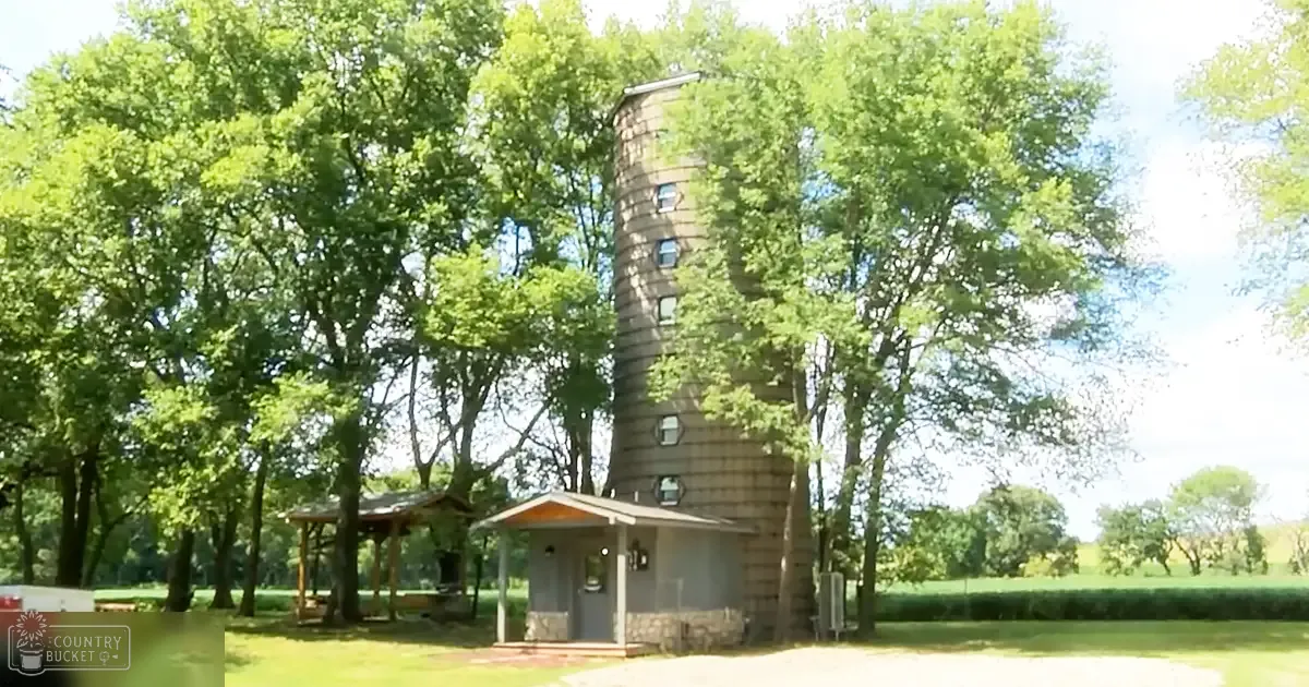 Rural scene with a silo-like tower, small structures, trees, and a white vehicle under clear daylight.