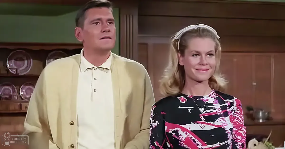 Adult male in a light yellow cardigan over a white collared shirt, standing with an adult female in a black dress with pink and white patterns, in a kitchen setting with wooden cabinets and daylight lighting.