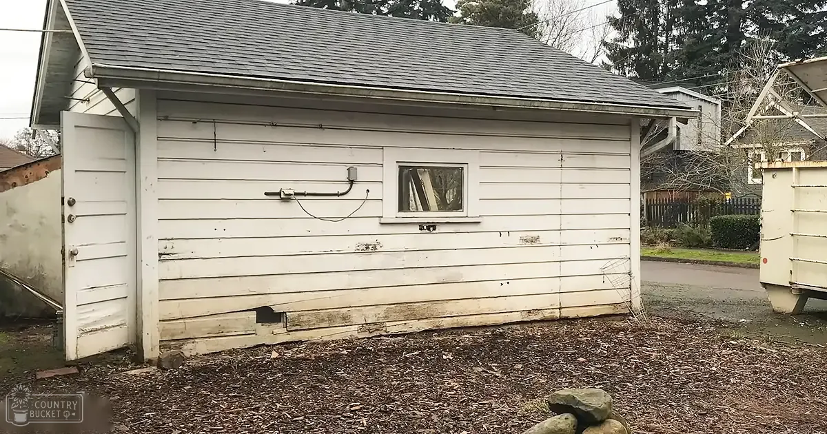 Small white shed with partially open window, metal pipe and hose attached to wall, mulch-covered ground, outdoor residential area with bare trees suggesting late fall or early spring, overcast daytime light