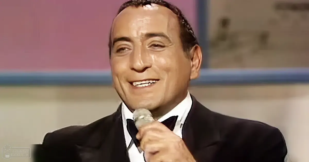 A middle-aged male singer in a black tuxedo with a white shirt and bow tie, holding a microphone on stage or in a studio setting.