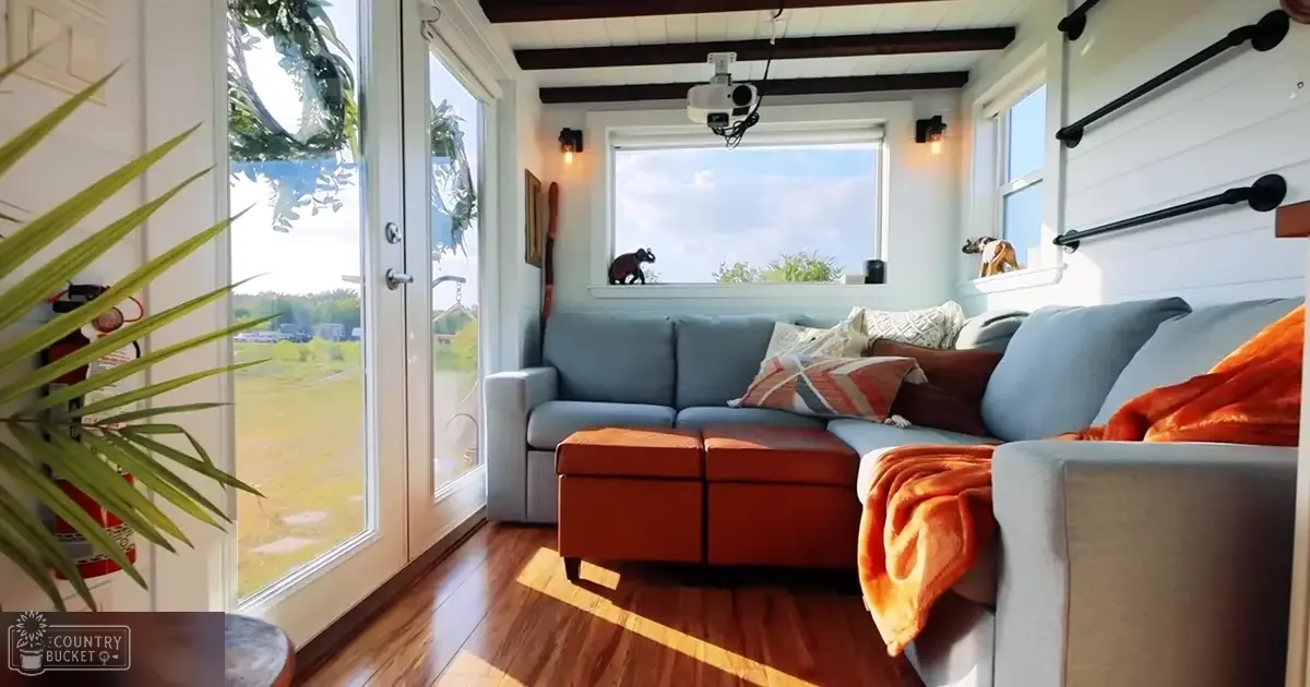 A cozy modern living room scene with a light gray sectional sofa, orange throw pillows, white walls, dark wood beams, and lush greenery outside the large windows during daytime.