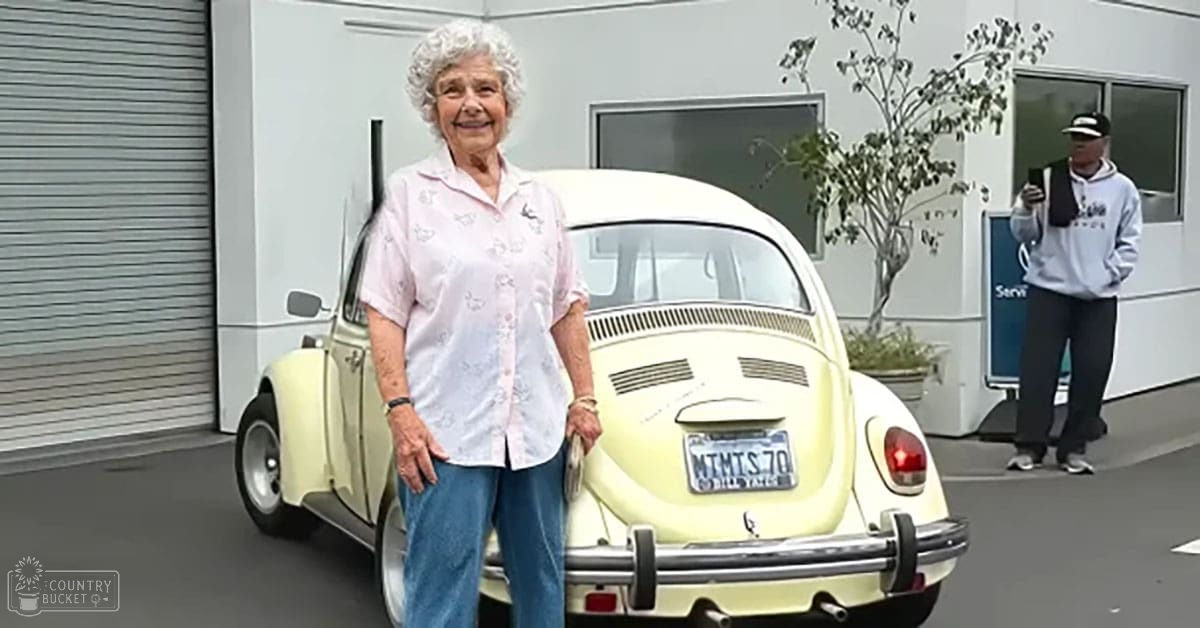 An older adult female wearing a light pink button-up shirt with small floral patterns stands next to a vintage Volkswagen Beetle at a service center, holding hands with an adult male in a light gray hoodie and black cap.