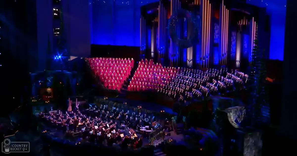 A choir dressed in red robes performs on a stage within an ornate concert hall under blue and purple stage lighting. Rows of seating for both the orchestra and the audience are visible, creating a formal atmosphere.