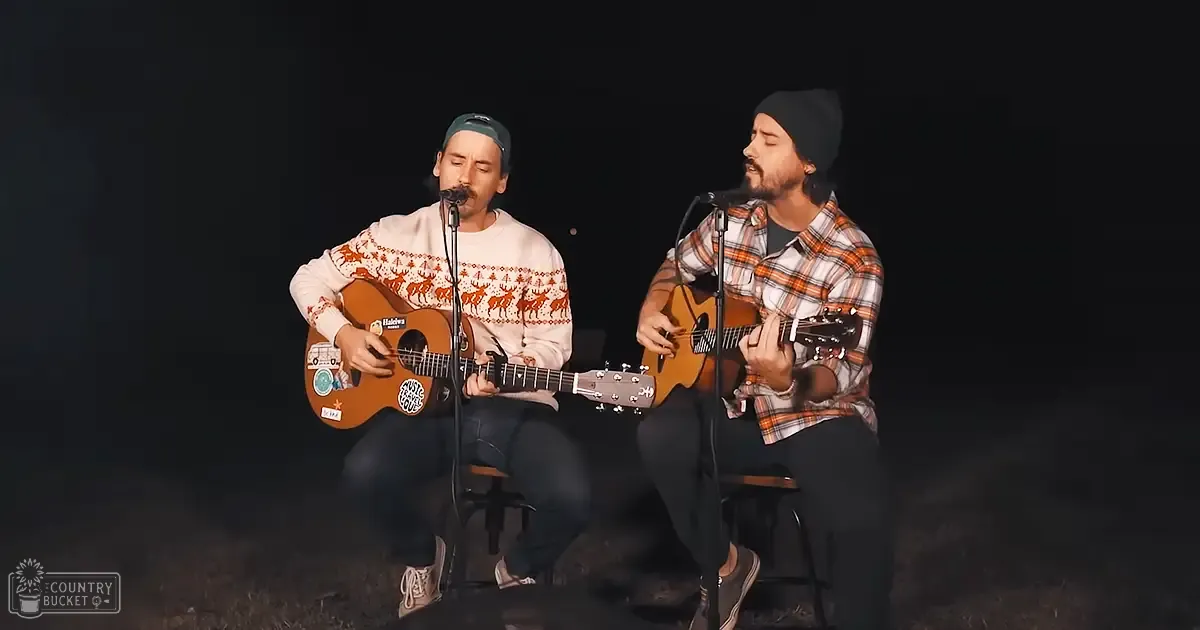 Two young adult male musicians performing outdoors at night, one wearing a white sweater with an orange reindeer pattern and playing an acoustic guitar decorated with stickers; the other in a plaid shirt of shades of orange, brown, and white. They sit on small stools against a dark backdrop under focused stage lighting.