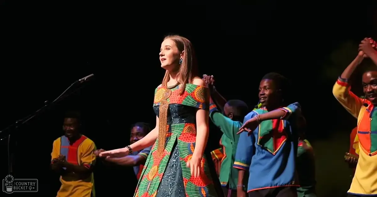 Young adult female in vibrant African-inspired performance attire, surrounded by male dancers in colorful traditional outfits on a stage with dark curtains and focused lighting.