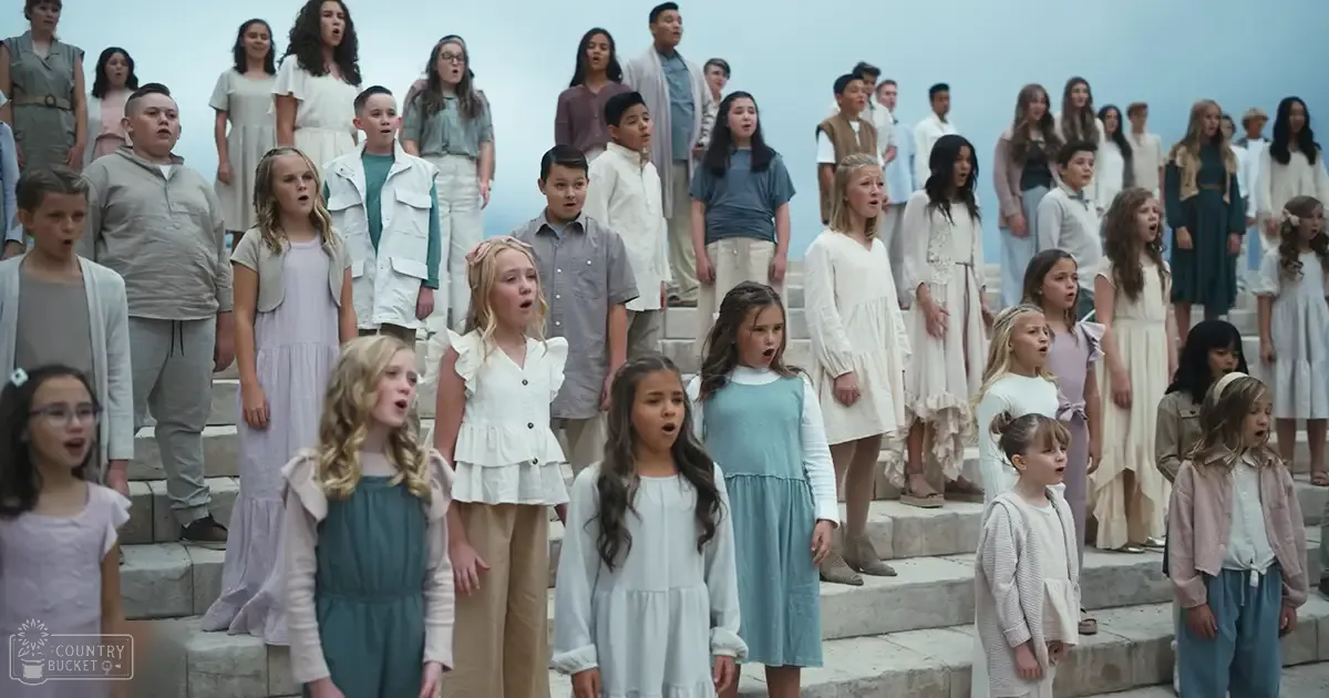 A group of children singing outdoors, each wearing a white or light-colored dress with ruffles.
