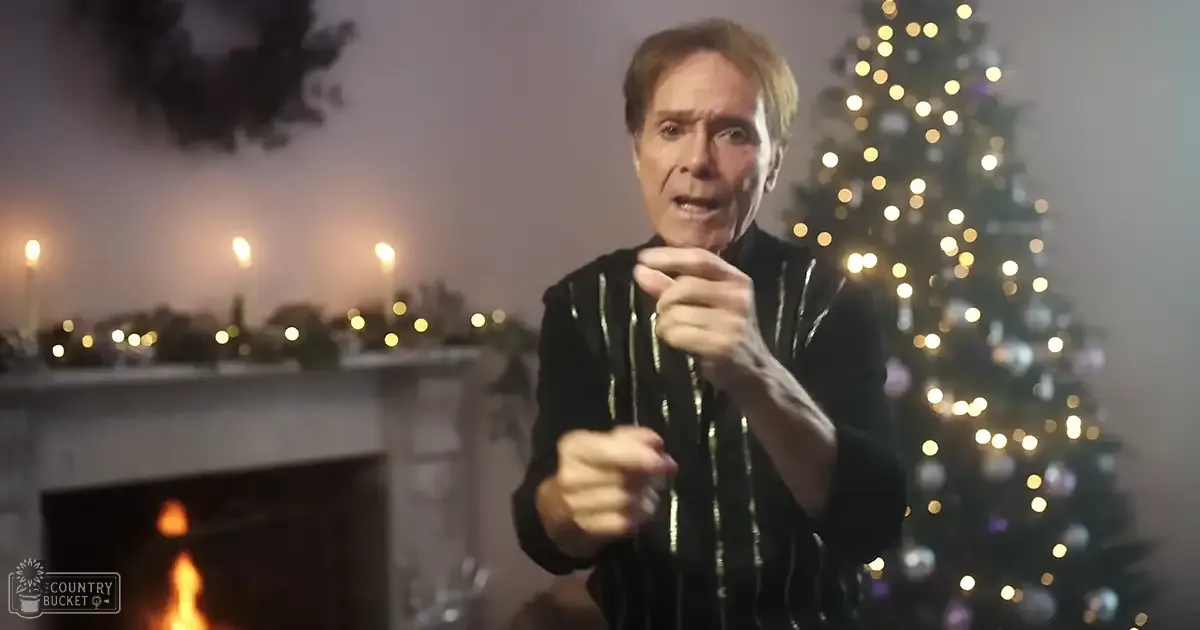 A middle-aged man wearing a black jacket with gold stripes stands indoors, surrounded by Christmas decorations including a lit fireplace and a decorated tree.