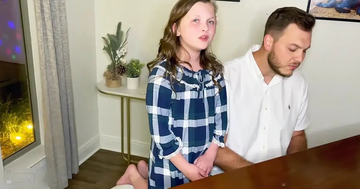 Child wearing a blue and white plaid dress with lace trim sits at a wooden table indoors, accompanied by an adult male in a white polo shirt; light gray or beige curtains hang near the window.